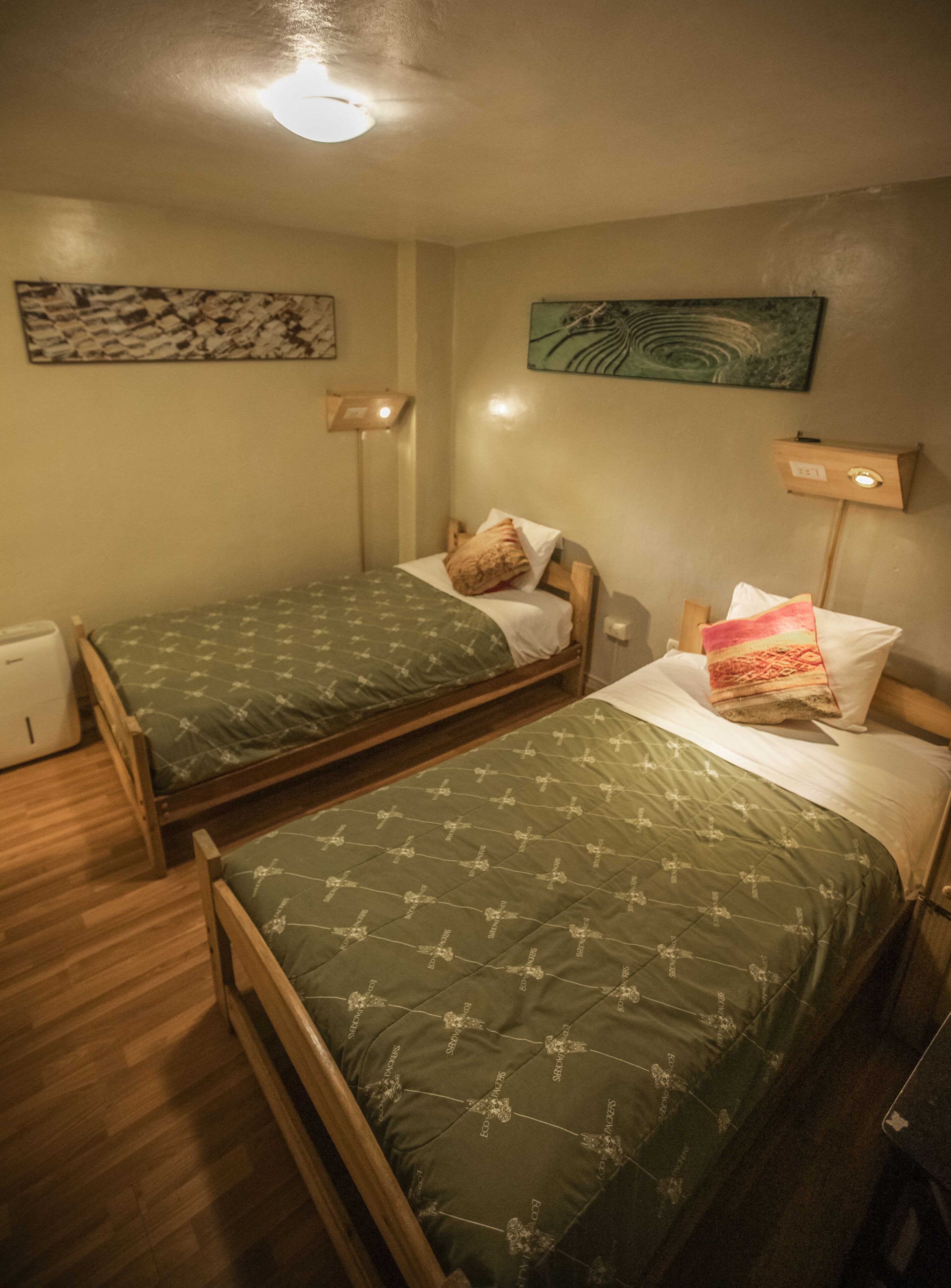 Une chambre avec deux lits simples, des têtes de lit en bois, des couvre-lits verts à motifs croisés, une petite table de chevet avec une lampe et un luminaire fixé au mur.