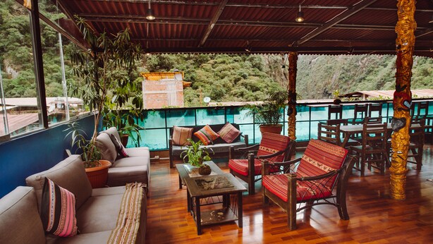 A spacious seating area with sofas, chairs, and a wooden table, surrounded by large windows offering a view of a lush hillside. - Nativus Hostel Machu Picchu (Machu Picchu)