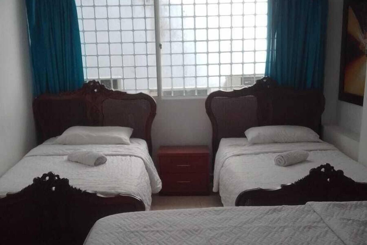 Una habitación de hotel con dos camas individuales, una cama más grande, una ventana con cortinas y un armario pequeño.