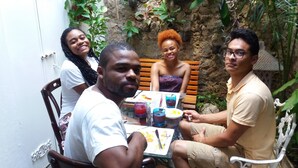 Un groupe de personnes est assis à une table à l’extérieur, devant des boissons et des assiettes de nourriture, entouré de verdure et d’un mur de pierre.