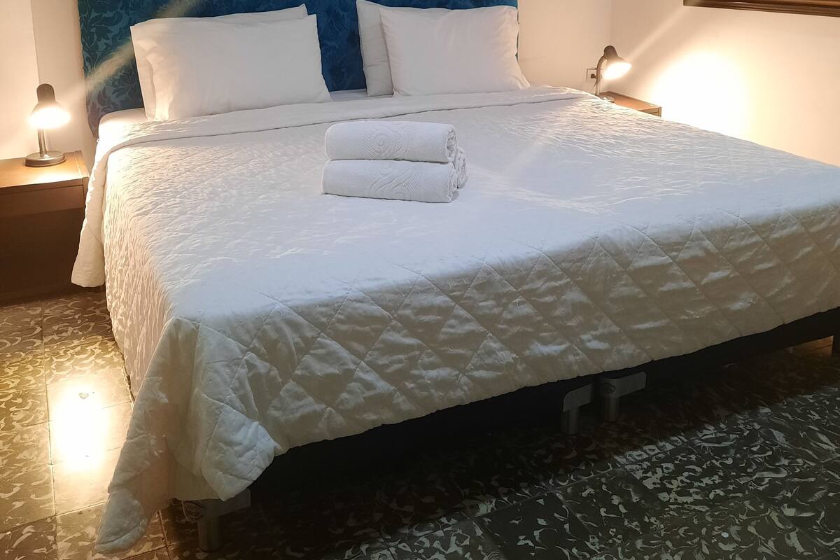 Una cama bien tendida con sábanas blancas y una toalla enrollada, un cabecero tapizado en azul y un cuadro en la pared.