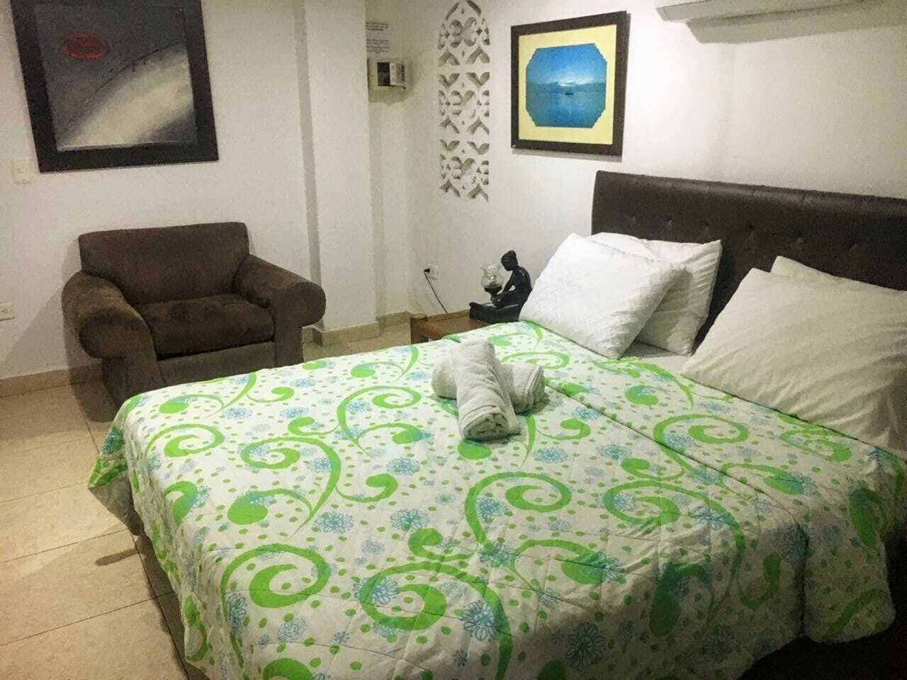 Un lit bien fait, recouvert d’une couvre-lit à motifs verts et blancs, un fauteuil marron et une petite statue posée sur une table de chevet.