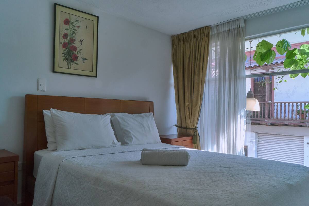 Una cama bien hecha con sábanas blancas, cabecera de madera y una mesita de noche con lámpara.