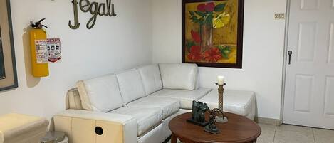 A living room with a white leather sofa, a wooden coffee table with a candle, a fire extinguisher, and a painting of flowers on the wall.