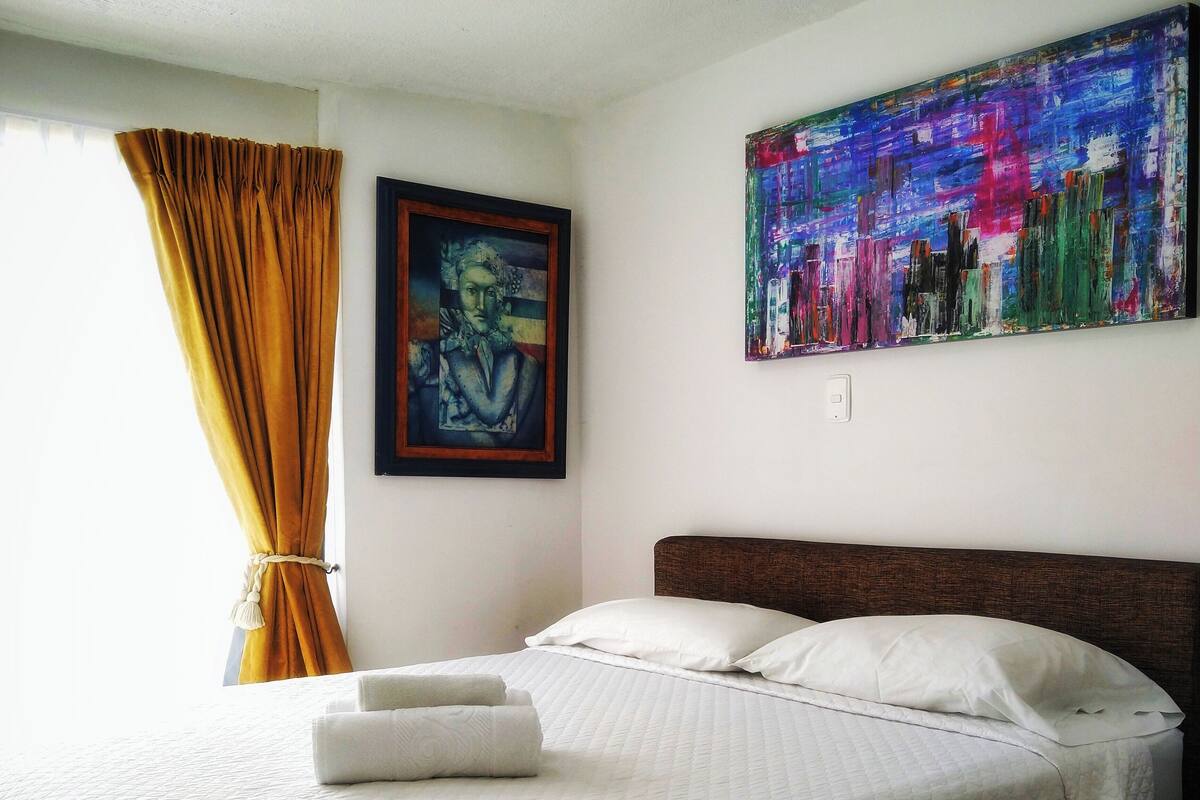 Una cama bien hecha con ropa blanca, un cuadro de paisaje urbano en la pared y una ventana con cortinas.