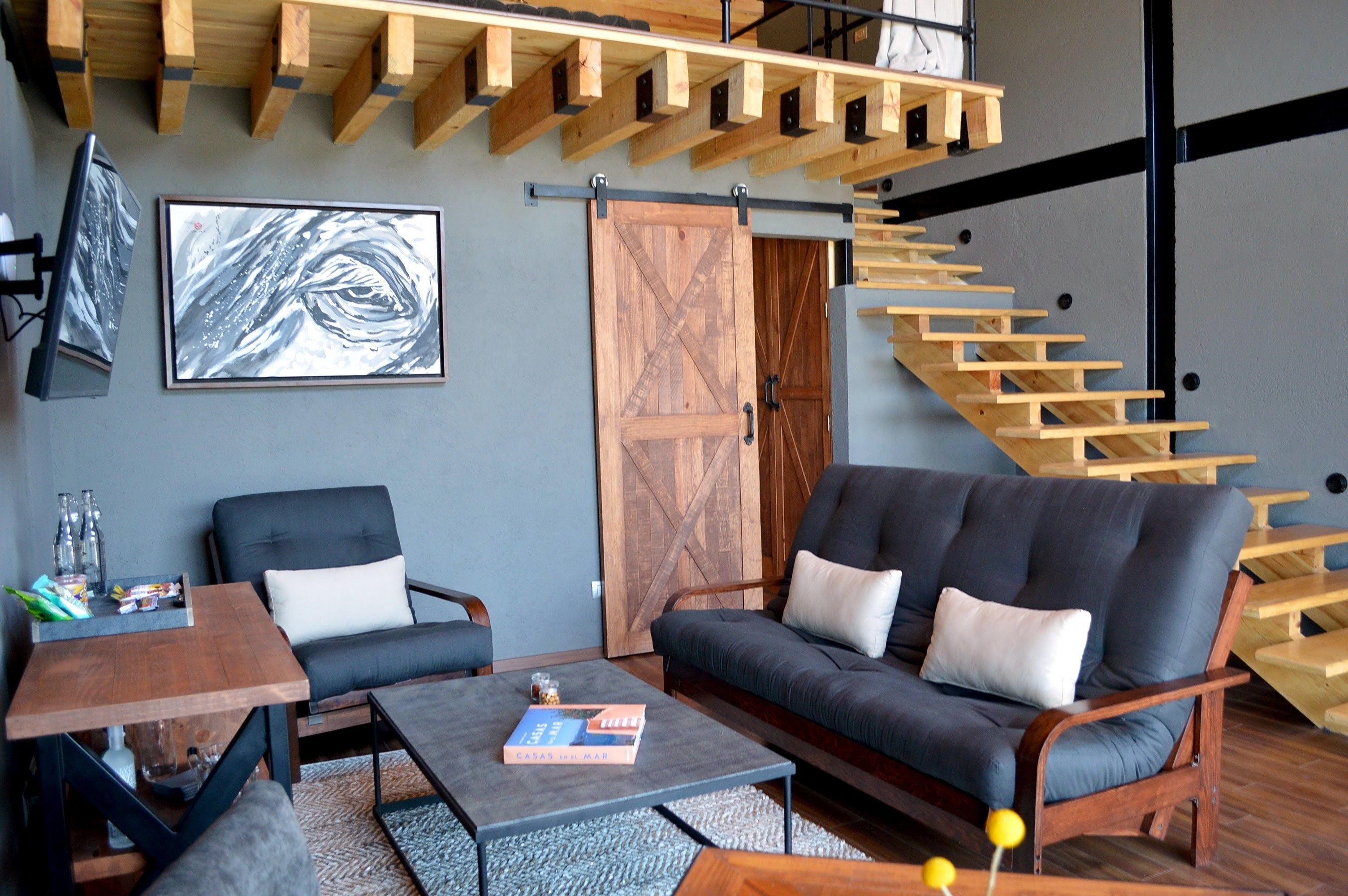 Una sala de estar moderna con un sofá, sillas, una mesa de centro y una estantería. Se ve una escalera de madera al fondo.