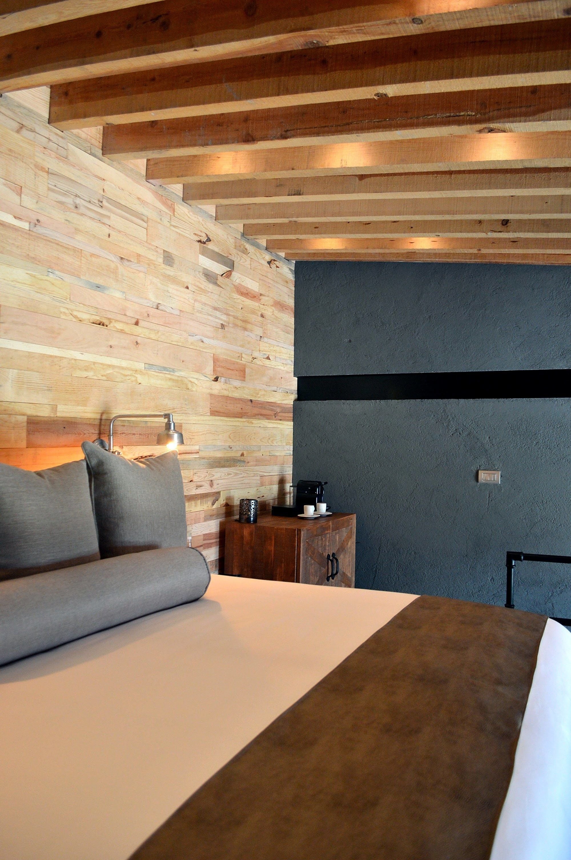 Quarto com parede de ripas de madeira, cama com colcha branca e marrom, sofá cinza, mesa de cabeceira com luminária e máquina de café.