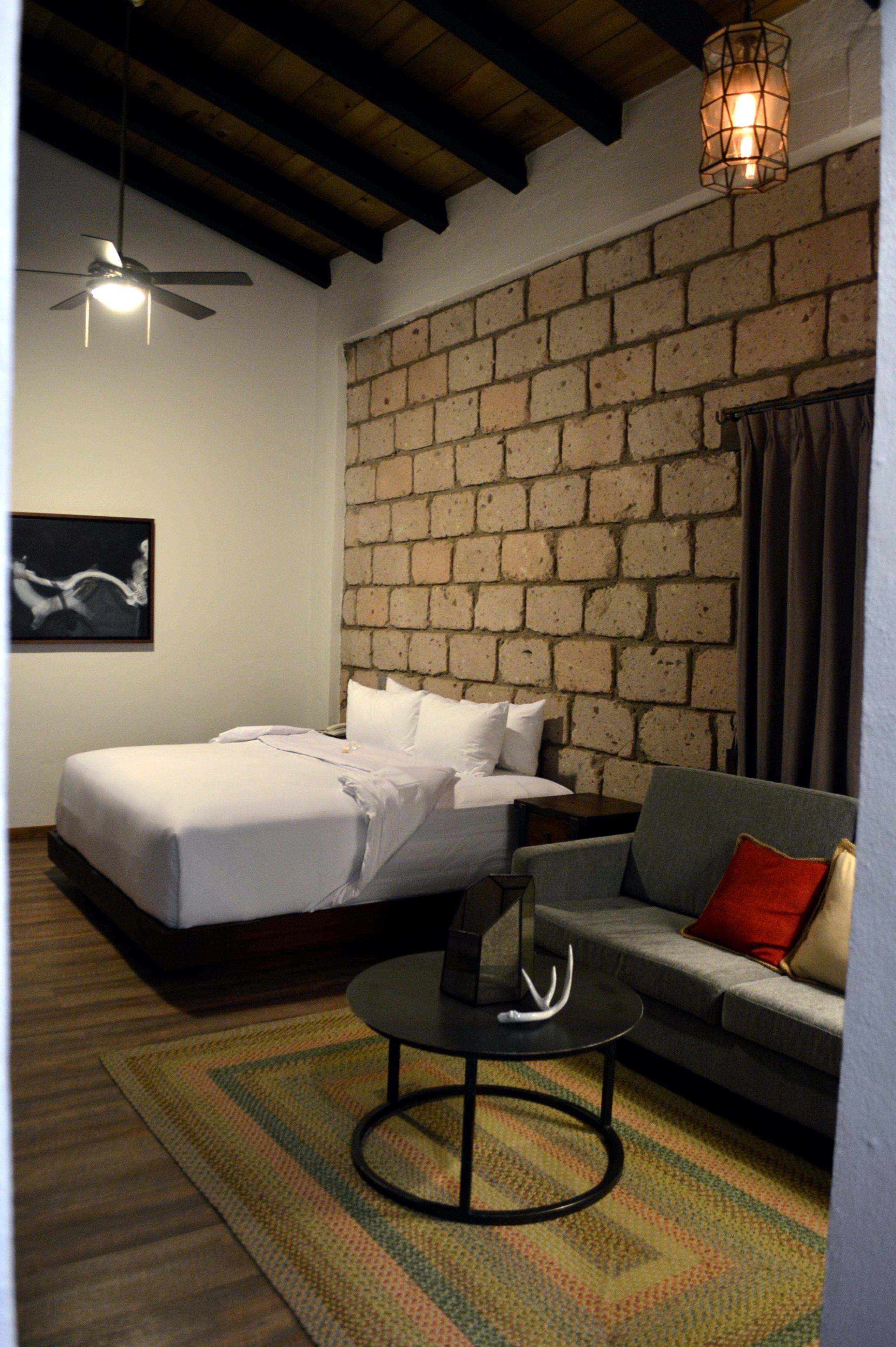 A bedroom with a bed, a sofa, a small table, a ceiling fan, and a brick wall.