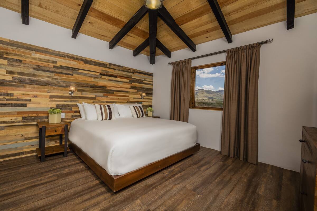 A bedroom with a wooden ceiling, a bed with white bedding, a nightstand with a lamp, and a window with curtains.