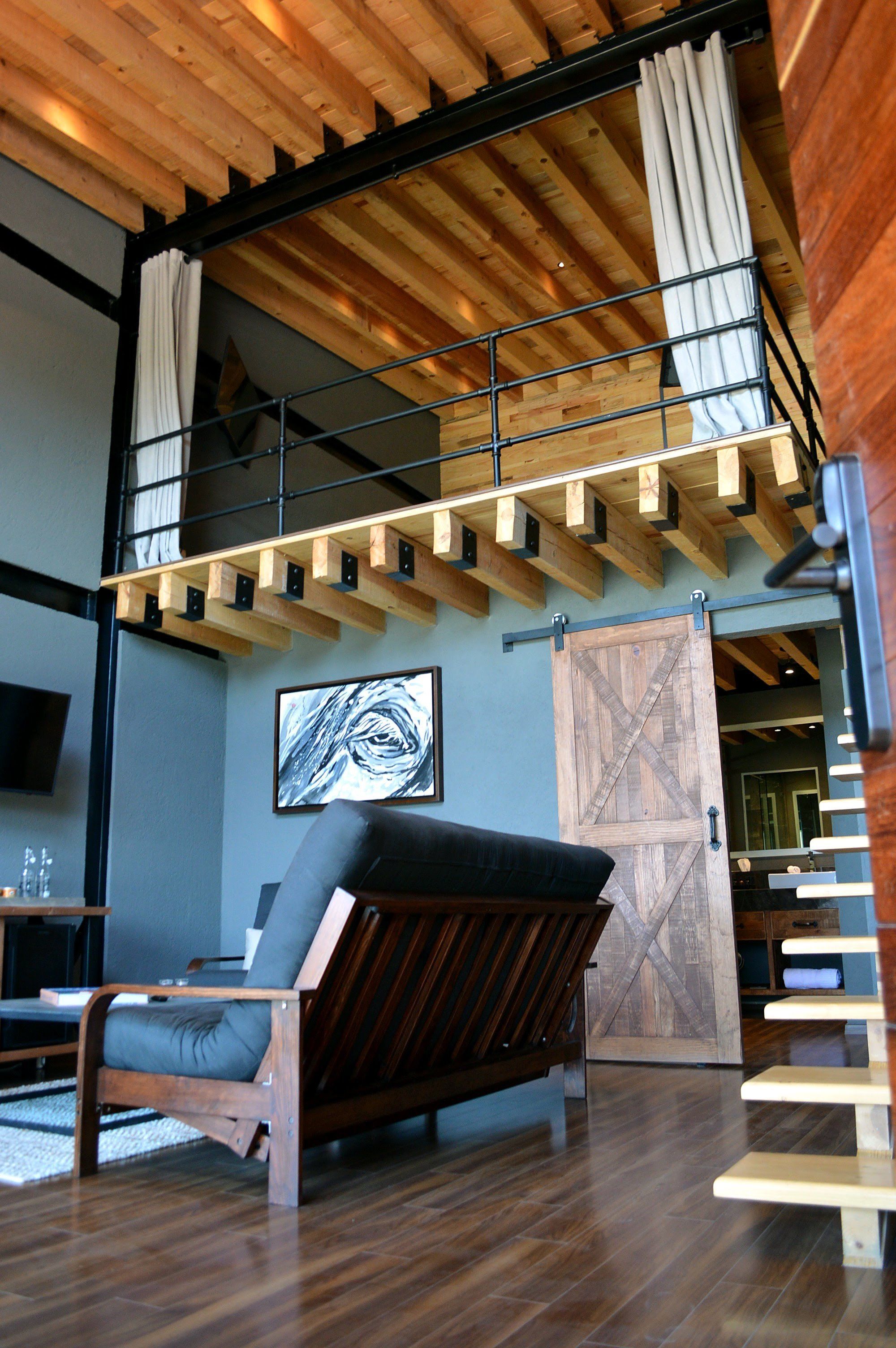 A loft-style room with a wooden ceiling, a large sliding barn door, a black sofa, and a staircase leading to an upper level.