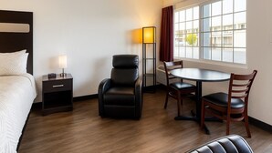 Habitación de hotel con cama, escritorio con lámpara, sillón de cuero, mesa de comedor con sillas y una ventana grande con cortinas.