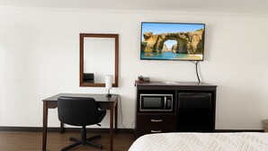 Habitación de hotel con cama, escritorio con silla, televisor montado en la pared, microondas y un mueble pequeño.
