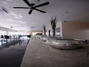 Um hall de hotel espaçoso, com ventiladores de teto, um balcão de recepção extenso e vista para o exterior através de amplas janelas.