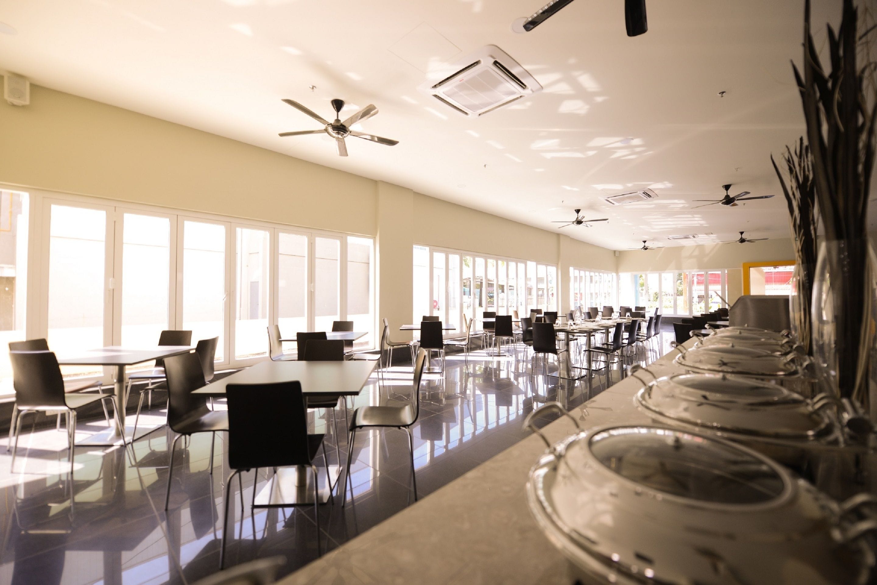 A spacious cafeteria with multiple tables and chairs, ceiling fans, and large windows allowing natural light.