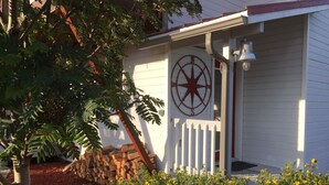 Une maison à l’extérieur blanc, ornée d’un symbole de boussole rouge, avec une clôture en bois peinte en blanc et un arbuste jaune en fleurs au premier plan.