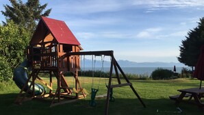 Une maisonnette en bois avec un toit rouge et un portique dans une cour verdoyante, offrant une vue sur les montagnes.