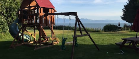 A wooden playhouse with a red roof and a swing set in a grassy yard with a view of mountains.