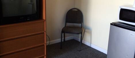 A room with a wooden TV stand, a small chair, a microwave, and a bed with patterned bedding.