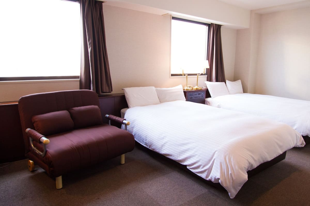 Une chambre d’hôtel avec deux lits, une chaise, une table de chevet et une fenêtre avec des rideaux.