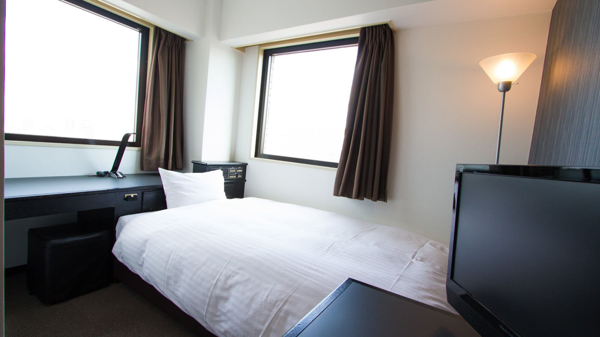 Een hotelkamer met een bed, een bureau, een stoel, een televisie en twee ramen met gordijnen.