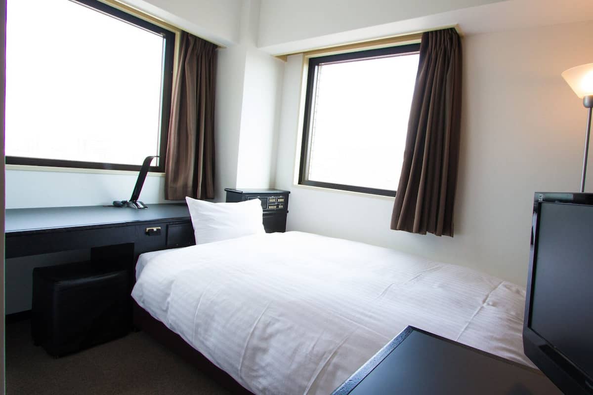 Une chambre d’hôtel équipée d’un lit, d’un bureau, d’une chaise, d’une télévision et de deux fenêtres avec des rideaux.