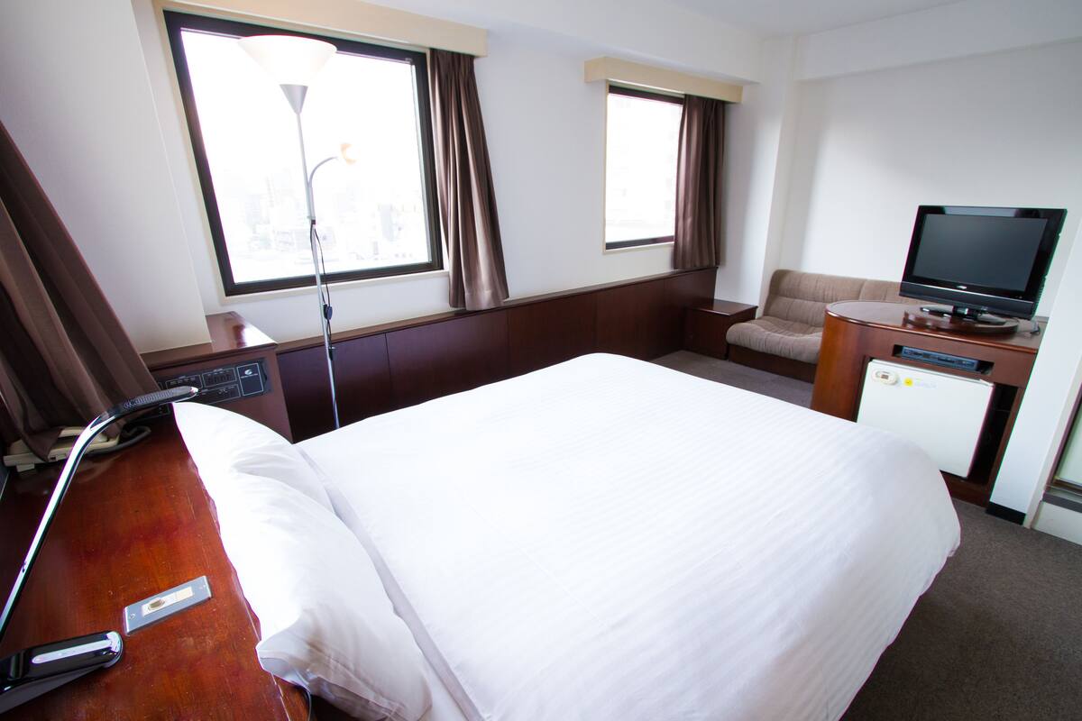 Une chambre d’hôtel avec un lit, un bureau avec un ordinateur, une télévision et deux fenêtres avec des rideaux.