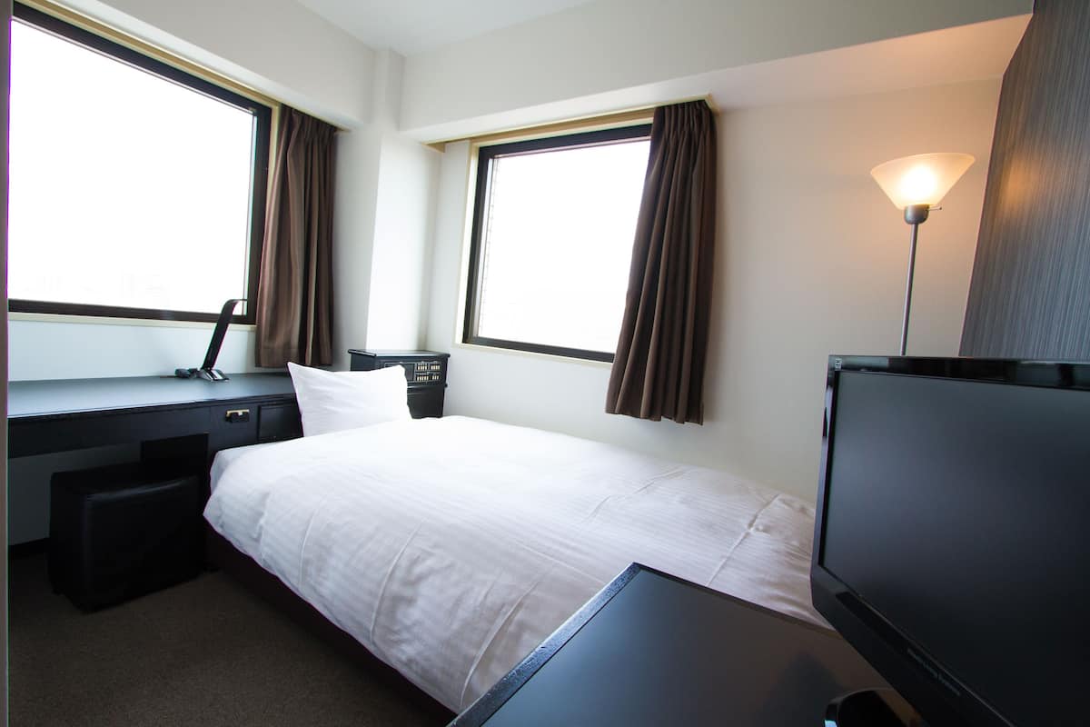Une chambre d’hôtel avec un lit, un bureau, une chaise, une télévision et deux fenêtres avec des rideaux.