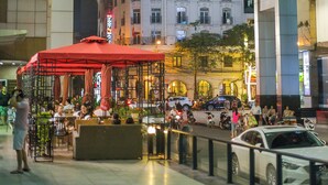 An outdoor cafe with tables and chairs, surrounded by a railing, in a city street with buildings and cars. - Midori Boutique Hotel (Hanoi)