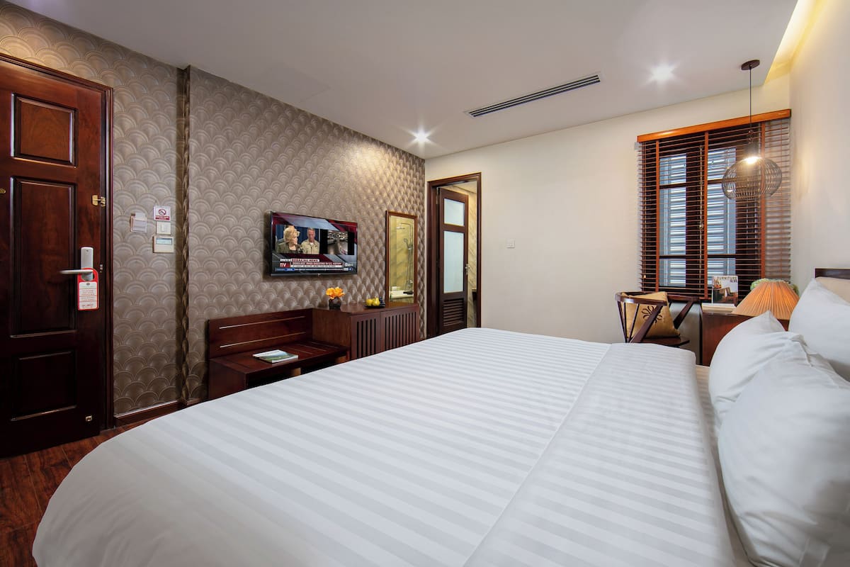 A hotel room with a large bed, a wooden wardrobe, a desk with a chair, a television, and a window with blinds.