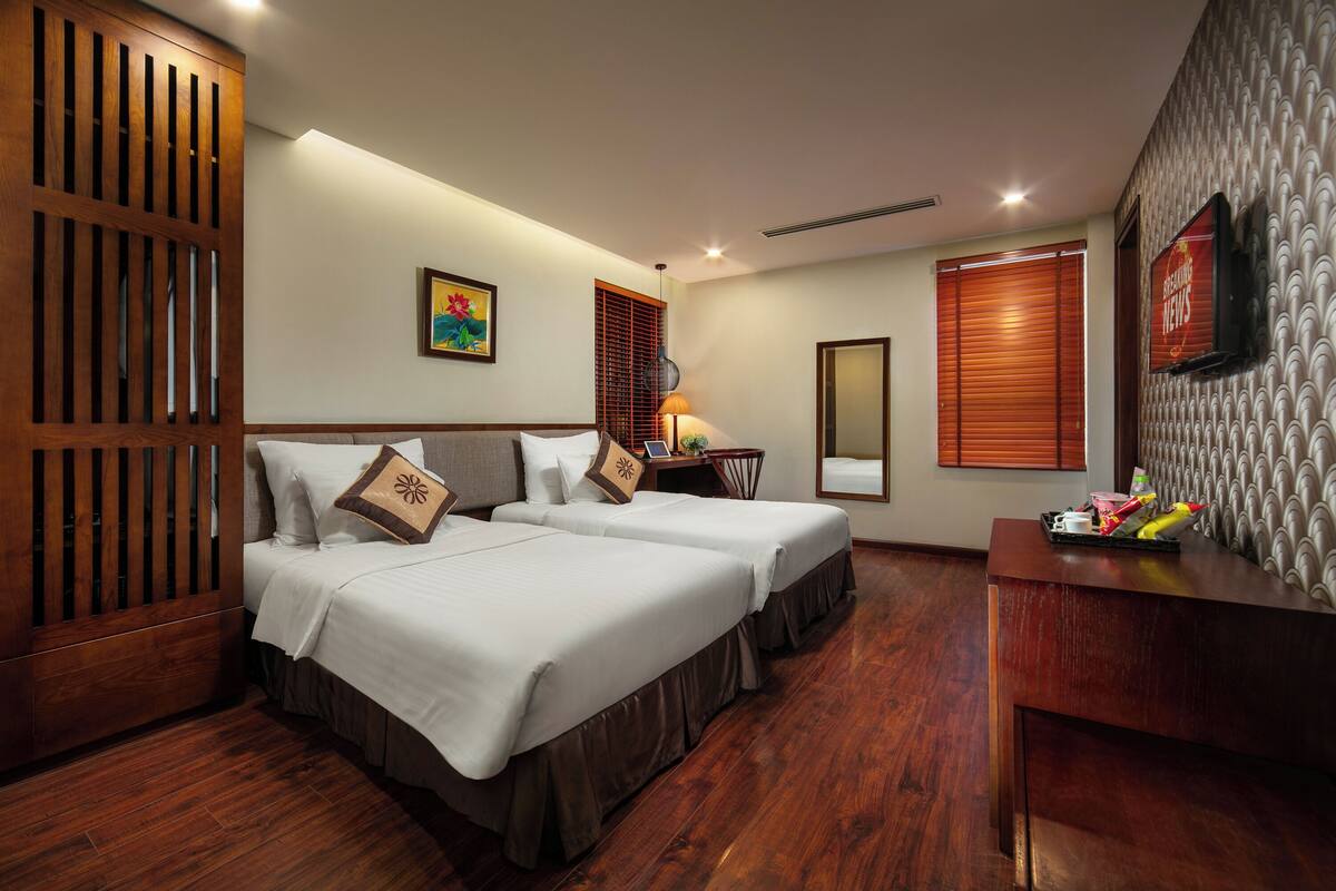 A hotel room with two beds, wooden flooring, a desk with a tray of items, a TV, and a decorative mirror.