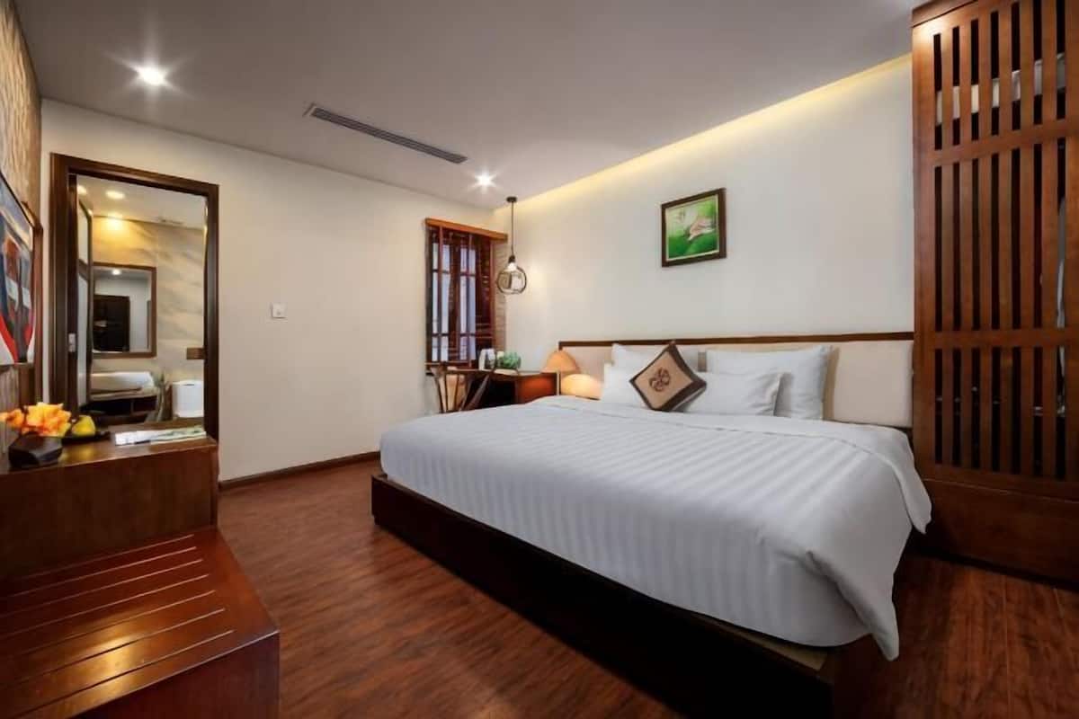 A hotel room with a large bed, wooden flooring, a desk with a chair, a mirror, and a wardrobe.