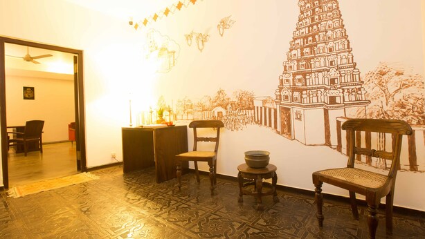 Um quarto com piso estampado, duas cadeiras de madeira, uma mesa pequena com uma tigela e um mural na parede retratando um templo.