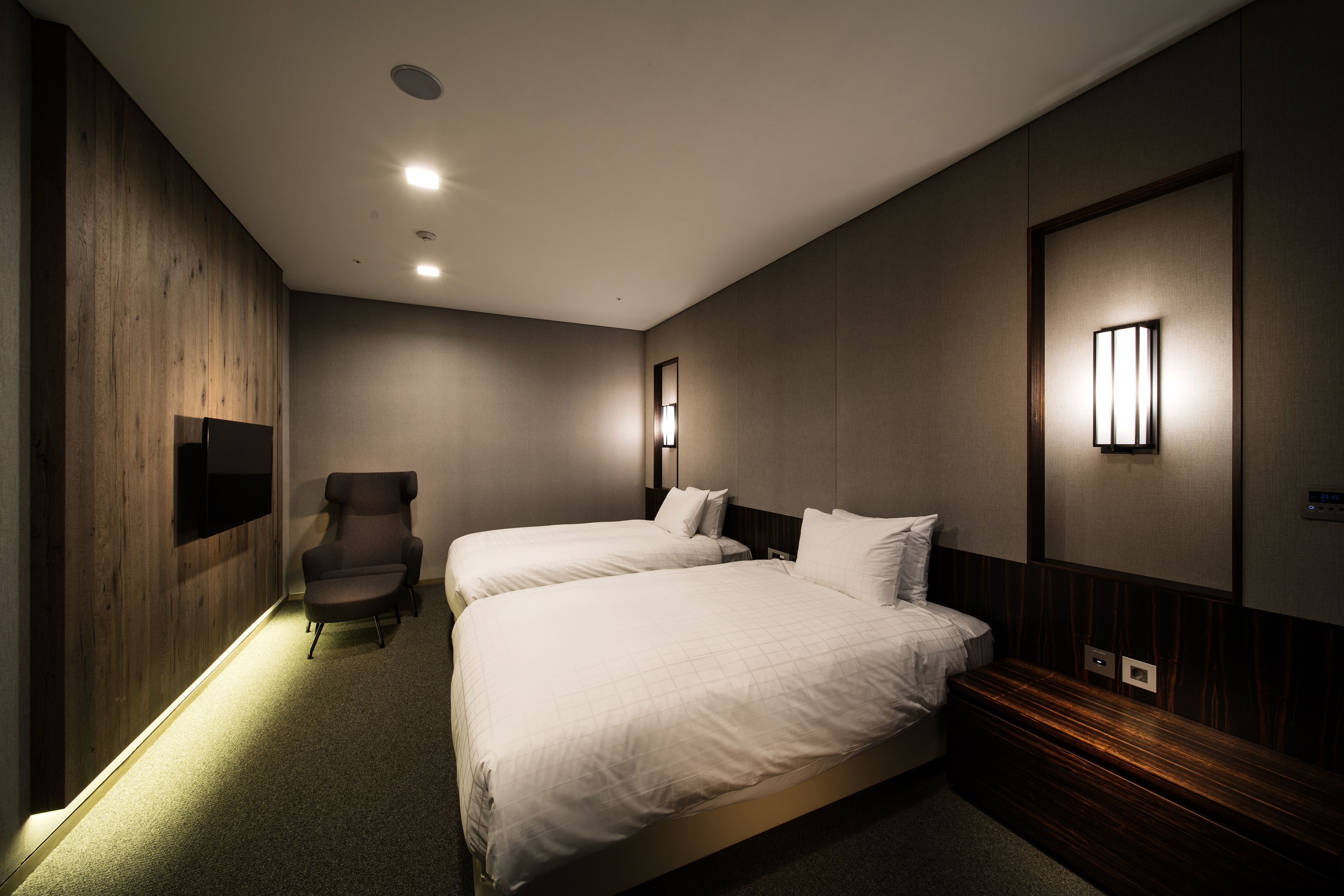 A hotel room with two beds, a chair, and a wall-mounted TV.