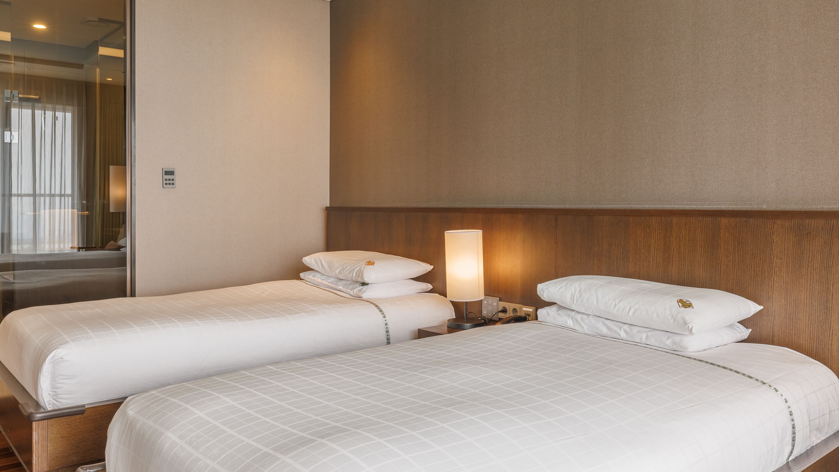 Habitación de hotel con dos camas, una mesita de noche con lámpara y un separador de vidrio que divide la habitación del baño.