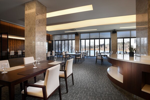 Un moderno vestíbulo de hotel con mostrador de recepción, varias mesas de comedor con sillas y grandes ventanales con vistas al exterior.