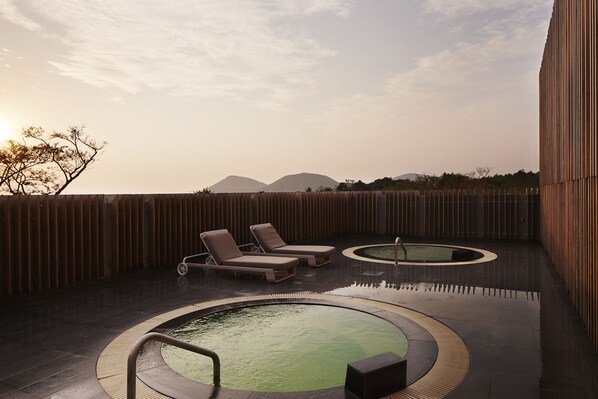 A rooftop pool area with lounge chairs and a hot tub, surrounded by a wooden fence and overlooking a mountainous landscape.