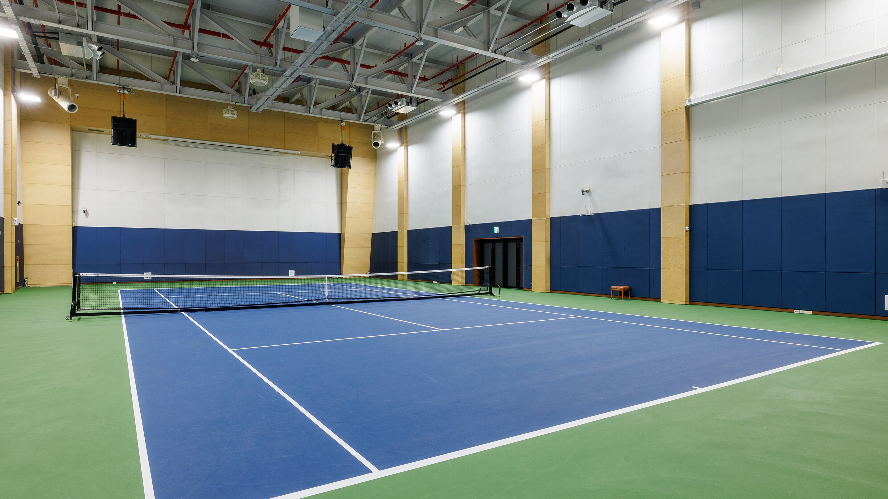 An indoor tennis court with a net, green and blue flooring, and a high ceiling with visible structural beams and lighting fixtures.