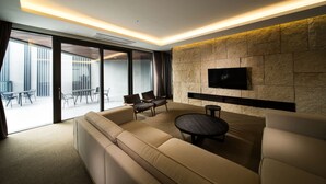 A modern living room with a sofa, chairs, a coffee table, and a flat-screen TV mounted on a textured wall. Large windows provide natural light and a view of an outdoor seating area. - The Annex (Seogwipo)