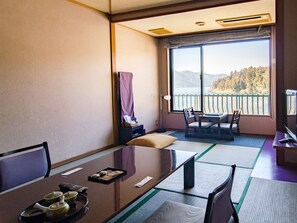 A room with a large window offering a view of a lake and mountains. There is a table with chairs, a TV, and a small table with cups and a tray. - Hotel Musashiya (Hakone)