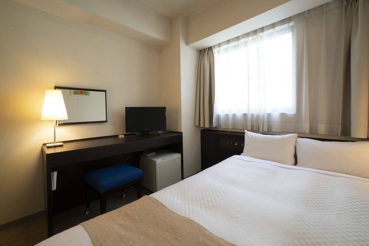 A hotel room with a bed, a desk with a computer, a small fridge, a lamp, and a window with curtains.