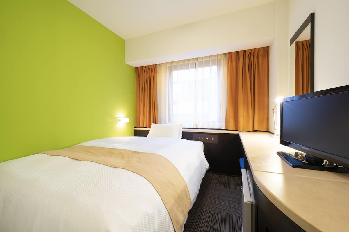 A hotel room with a bed, a desk with a computer, a TV, and a window with curtains.