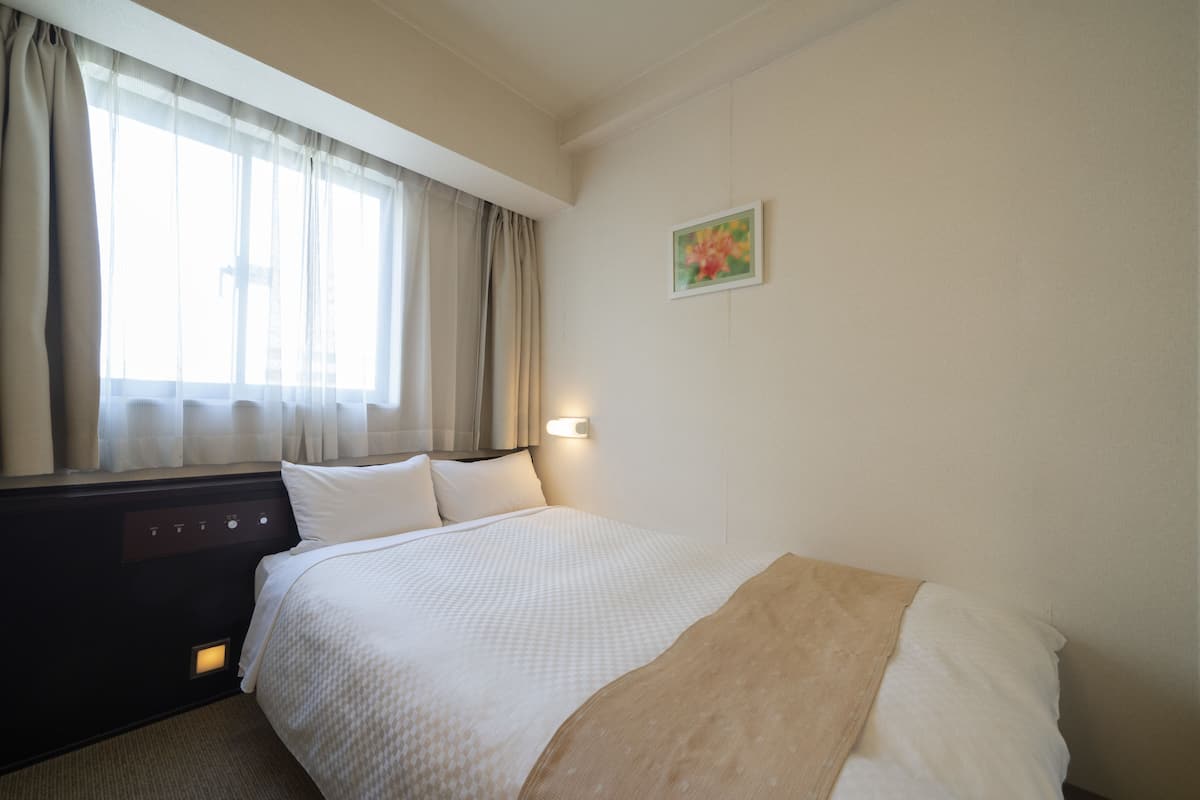 A hotel room with a bed, curtains, a painting, and a control panel.