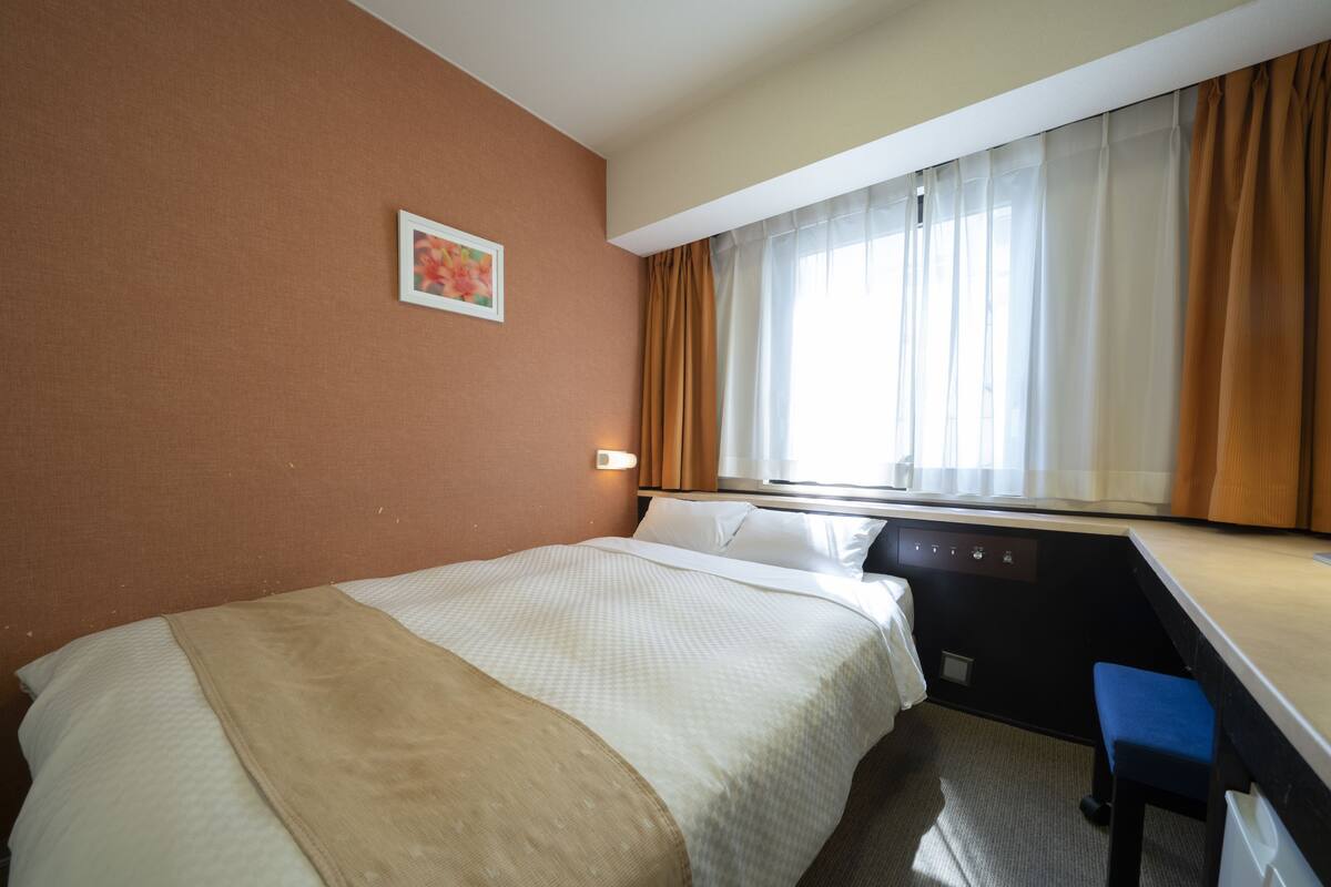 A hotel room with a bed, a desk, a chair, a window with curtains, and a wall-mounted light.