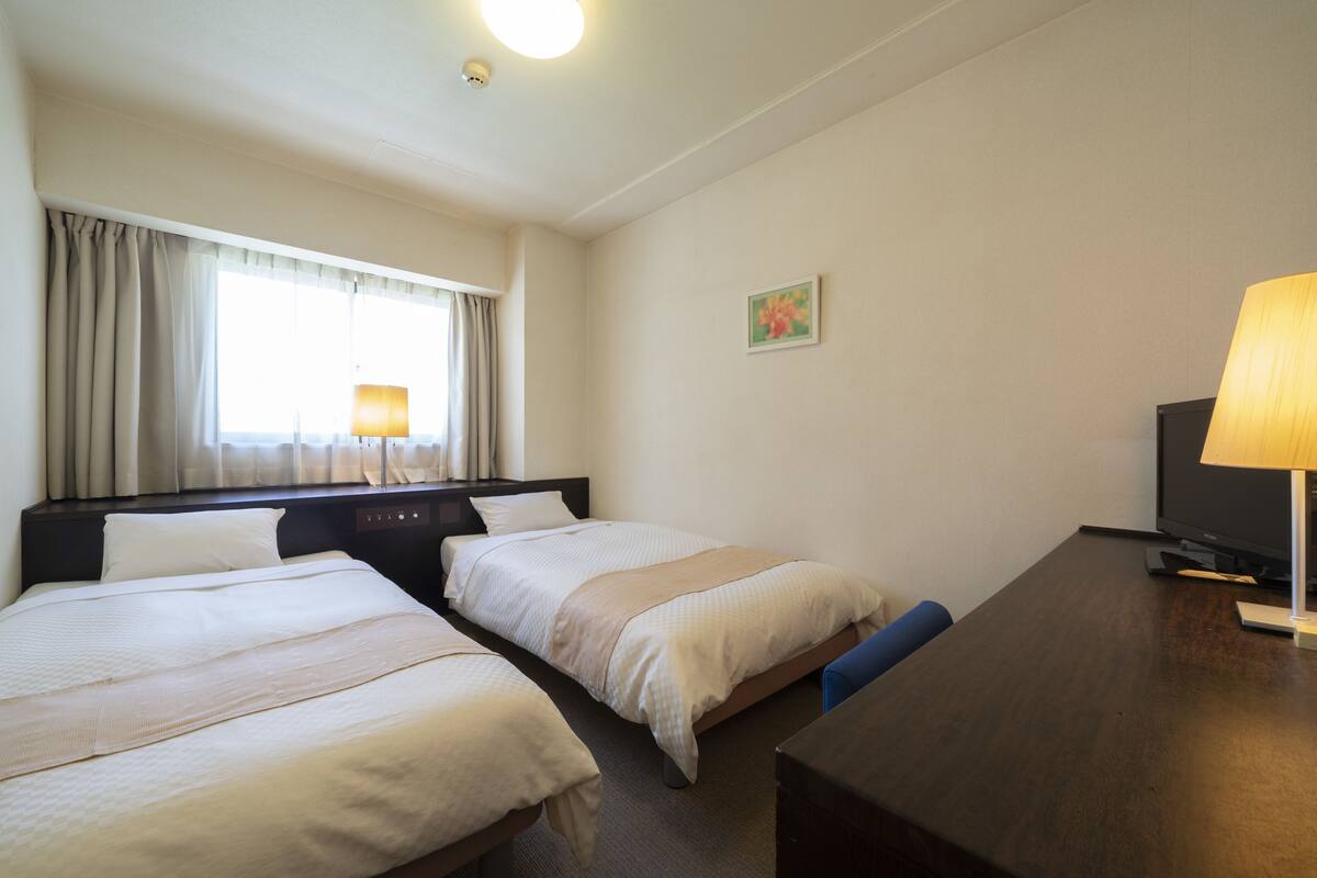 A hotel room with two beds, a desk, a television, and a window with curtains.