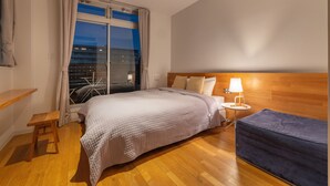 A hotel room with a large bed, a wooden headboard, a nightstand with a lamp, a window with curtains, and a view of the city at night. - UNPLAN Kagurazaka - Hostel (Tokyo)