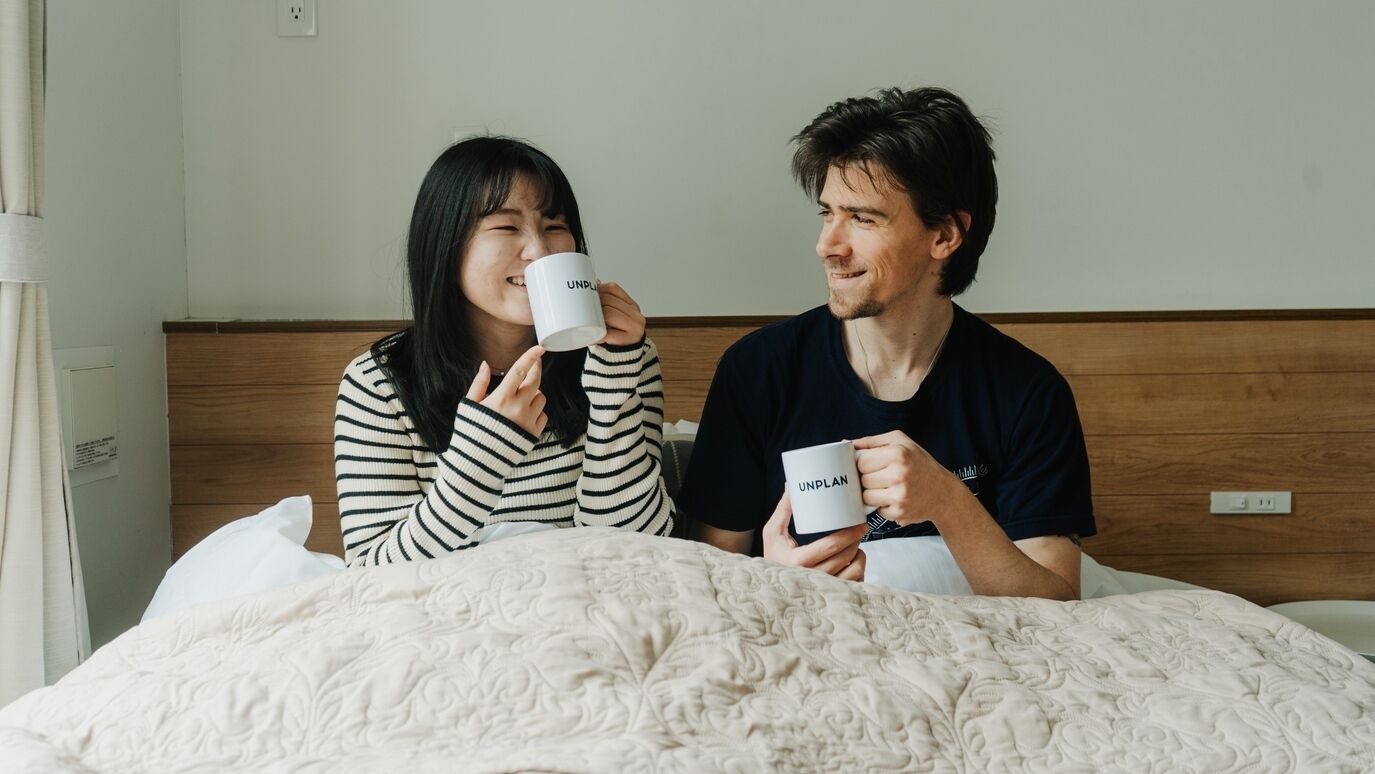 Two people sitting on a bed with a wooden headboard, holding mugs with text on them.