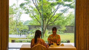 A room with a table and chairs, a window with curtains, and a view of a garden with a tree and a fence.