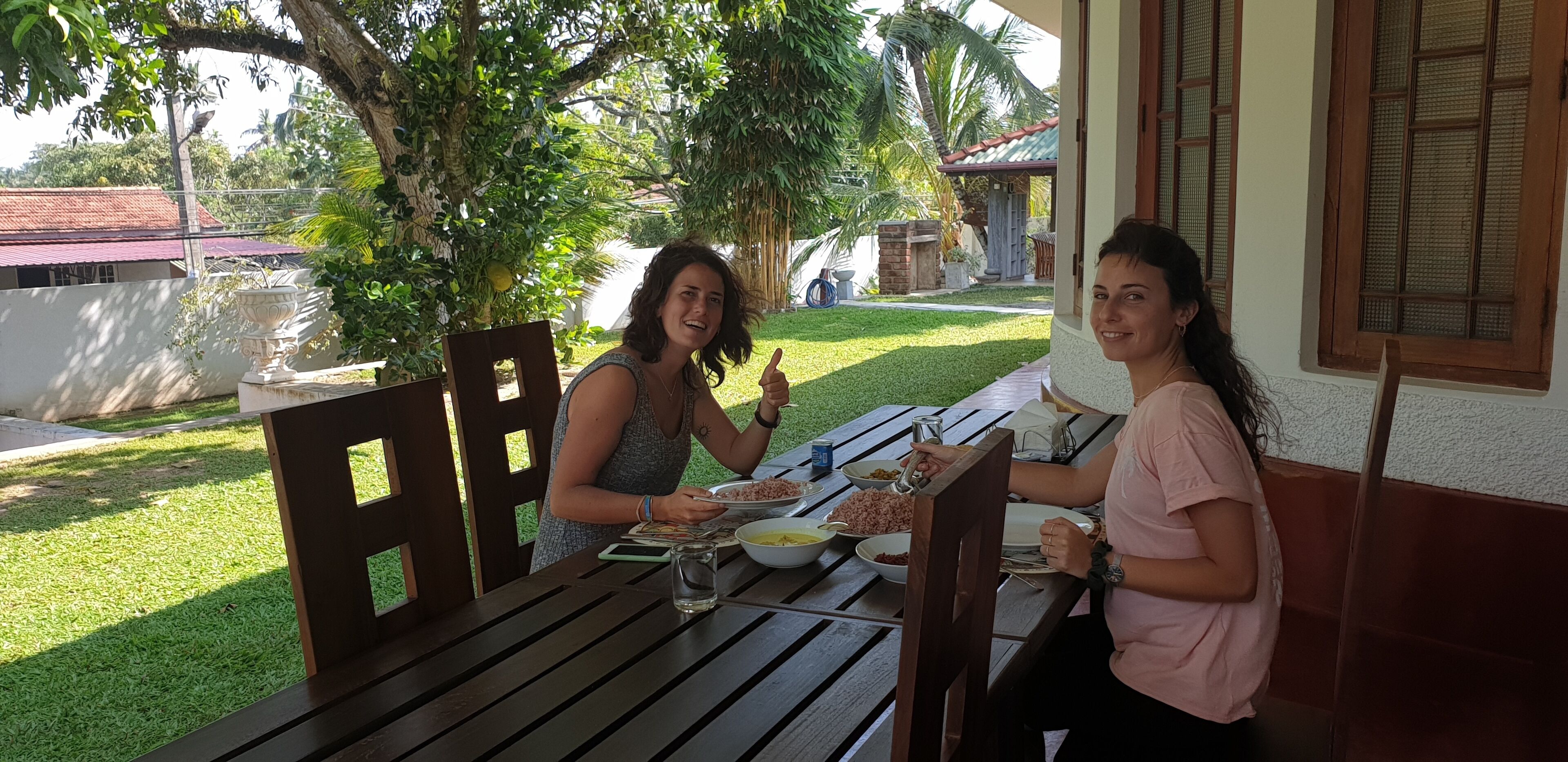 Two people dining at an outdoor table with a view of a garden and a building in the background.