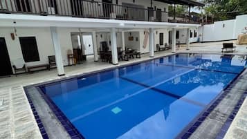 A swimming pool with a tiled deck, a covered seating area, and a building with multiple balconies.