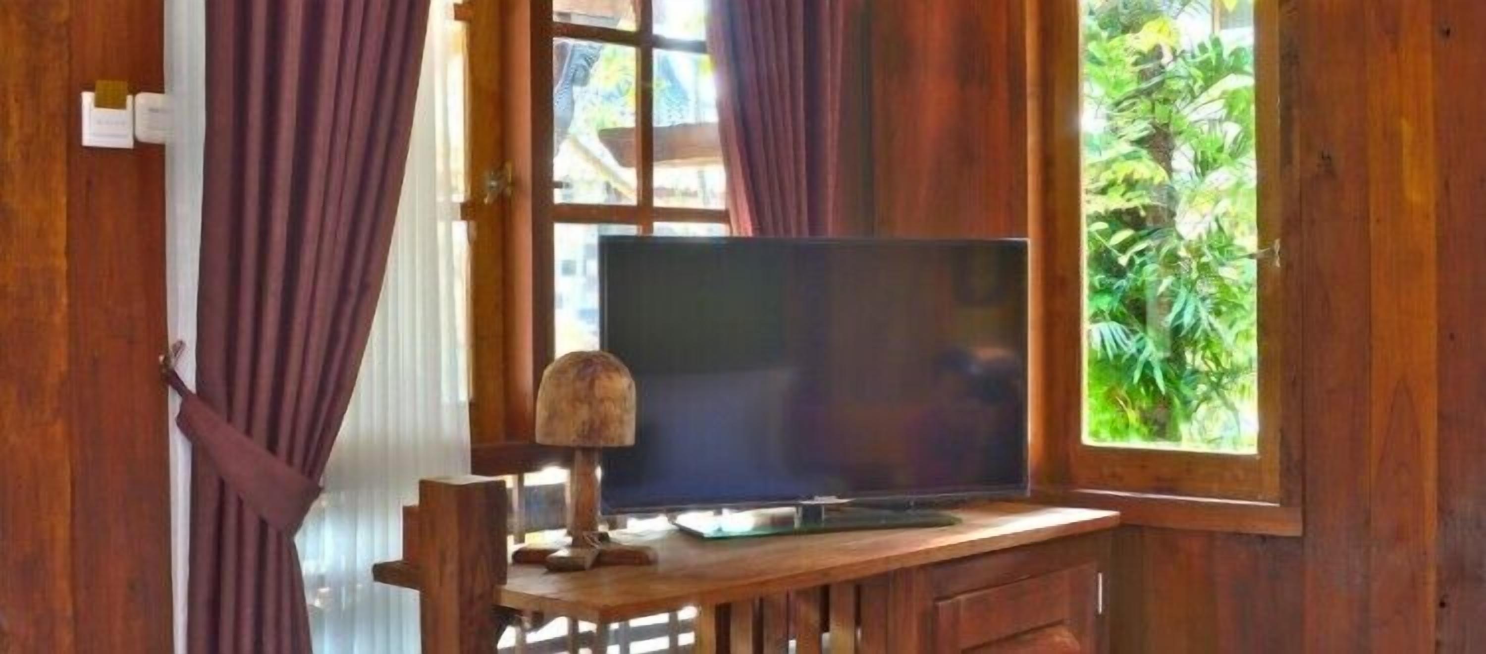 A wooden desk with a television, curtains, and a window with a view of greenery.